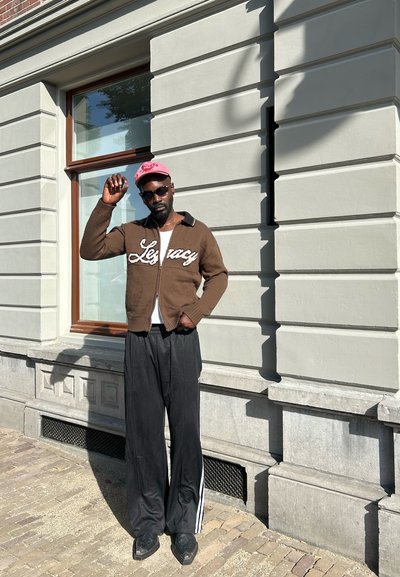 Suéter marrón con cierre y texto "Legacy", pantalones anchos negros con rayas blancas, zapatos negros y una gorra rosa; de pie contra un edificio.
