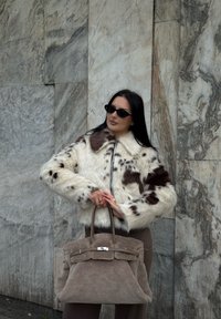 Kvinde iført en brun og hvid pelsjakke med solbriller, der holder en stor taupe tekstureret håndtaske mod en marmorvæg.