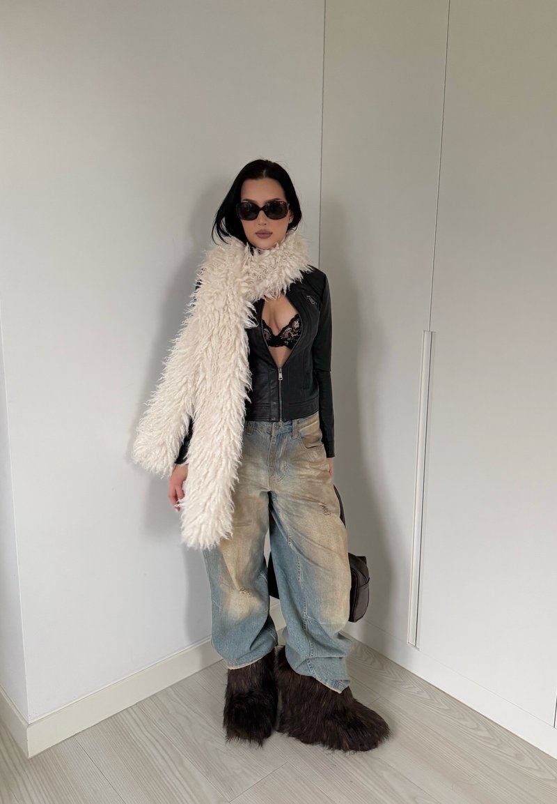 Veste en cuir noire, soutien-gorge en dentelle, jean bleu délavé oversize, écharpe blanche en fourrure douce, et bottes brunes en fourrure. Lunettes de soleil foncées et accessoires minimalistes.