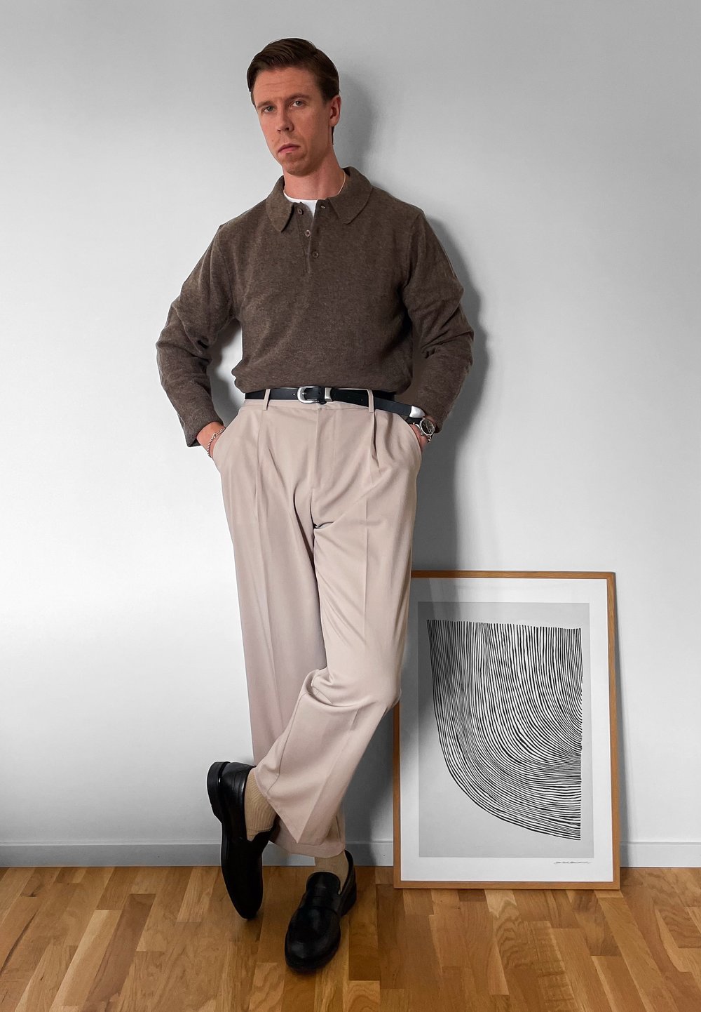 Polo din lână maro, pantaloni plisati bej, curea neagră și pantofi. Stând lângă o lucrare de artă abstractă în alb și negru, încadrată.