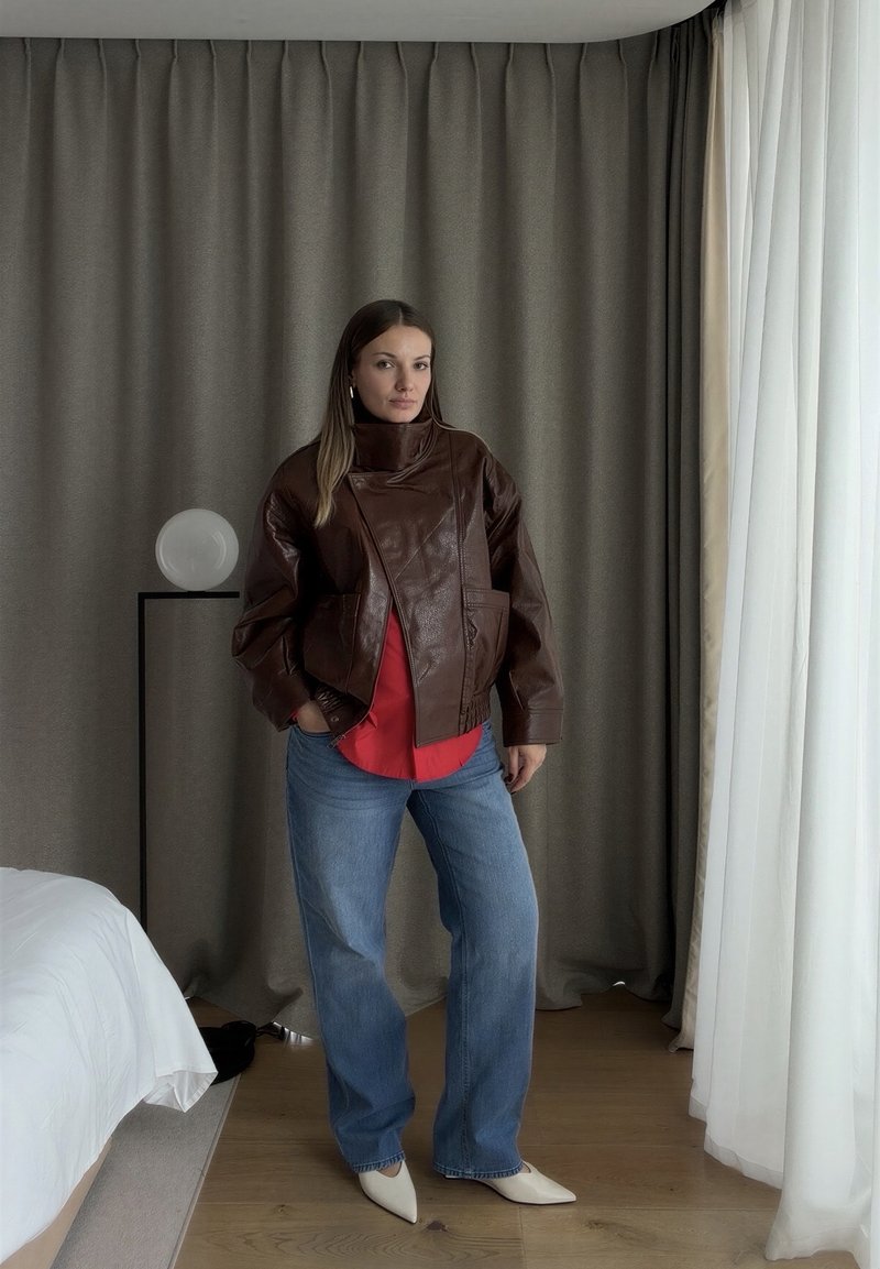 Une femme pose debout dans une chambre, portant une veste en cuir marron sur un haut rouge, un jean large et des chaussures pointues.