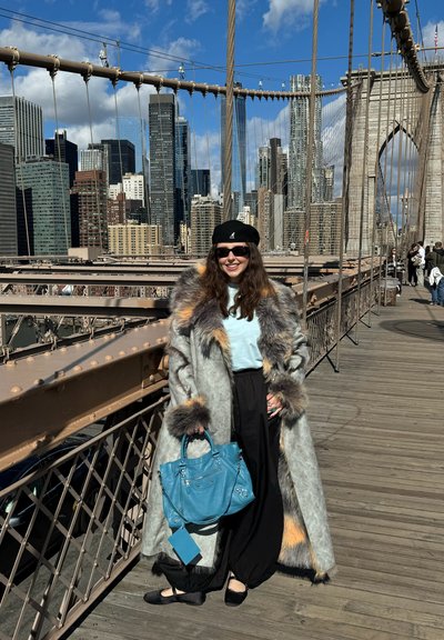 Mujer con abrigo largo con ribete de piel, pantalones negros, boina y gafas de sol sosteniendo un bolso azul en el Puente de Brooklyn con el horizonte de la ciudad detrás.