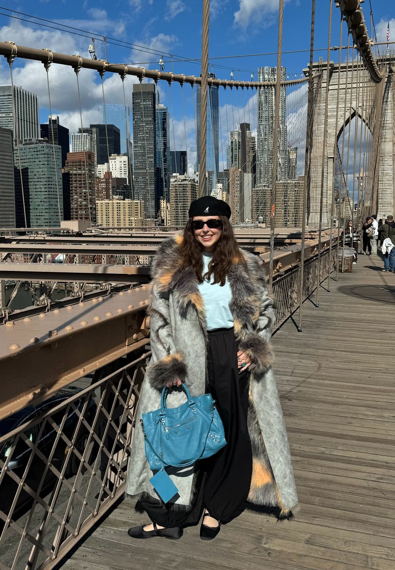 Femme portant un long manteau bordé de fourrure, un pantalon noir, un béret et des lunettes de soleil tenant un sac à main bleu sur le pont de Brooklyn avec la silhouette de la ville en arrière-plan.