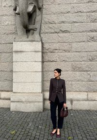 Vrouw in een donker jasje en broek, met een geweven handtas, staat op een klinkerstraat voor een stenen muur met een olifantbeeld.