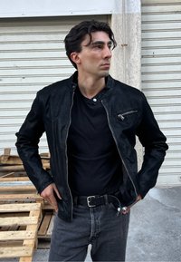 Melnas ādas jaka ar rāvējslēdzēja priekšpusi, ribotām apkaklēm un sānu kabatu. Valkāta virs melnas T-krekla un pelēkiem džinsiem. Vienkāršs fons.