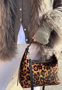 Main tenant un sac à main imprimé léopard, portant un pull gris duveteux et un cardigan marron boutonné sur un pantalon blanc.