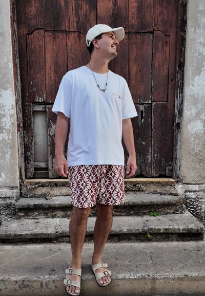 Joven con gorra blanca, camiseta blanca, pantalones cortos con estampado y sandalias beige, de pie sobre unos escalones de piedra frente a una puerta de madera envejecida.