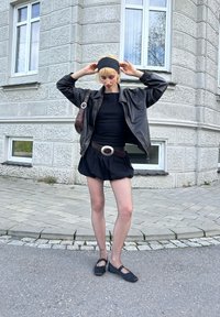 Μαύρο δερμάτινο μπουφάν τύπου bomber, μαύρη εφαρμοστή μπλούζα, μαύρη φουστάνα με φαρδιά κρόσι, καλσόν ψαθωτό και μαύρα φλατ παπούτσια. Σημαντική ασημένια αγκράφα ζώνης ως λεπτομέρεια.