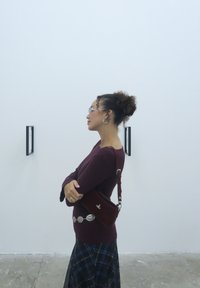 A woman with curly hair tied up, wearing glasses, a burgundy top, and a plaid skirt, stands with her arms crossed, admiring wall art.
