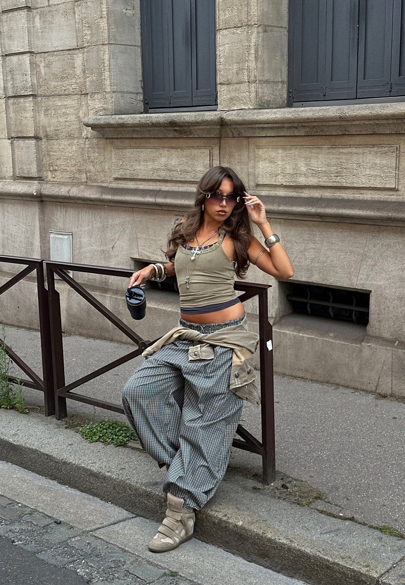 Jeune femme en pantalon ample et débardeur s'appuie sur une rambarde métallique sur le trottoir de la ville, tenant une boisson et ajustant ses lunettes de soleil.