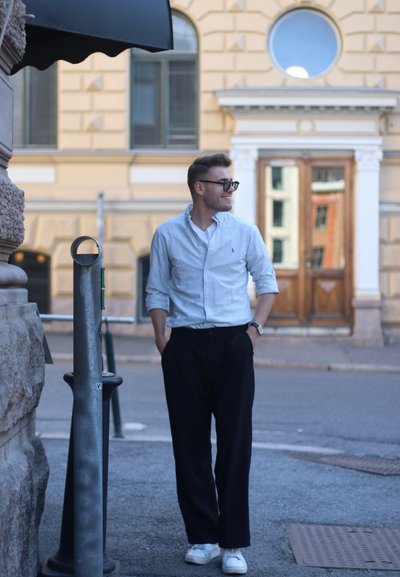 Camisa de botones a rayas azul claro, pantalones de pierna ancha oscuros, zapatillas blancas y gafas negras, de pie junto a una pared de piedra y un poste de metal.