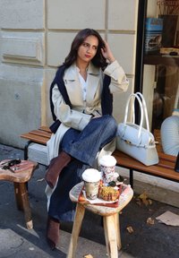 Hellbeiger Trenchcoat mit einem marineblauen Schal, blaue Jeans mit weitem Bein und braune spitze Stiefeletten. Hellgraue Handtasche und Kaffeetassen auf Holzschemeln.