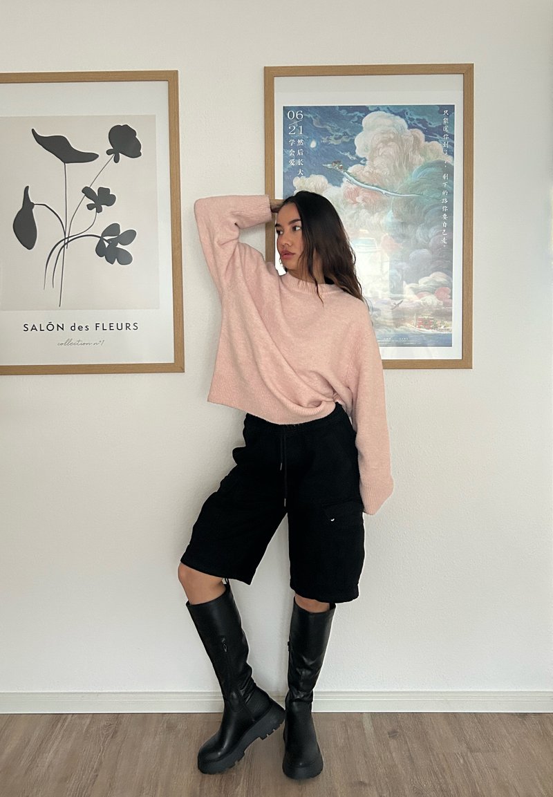 Suéter malhado rosa claro, calções de carga pretos até ao joelho e botas pretas até ao joelho. Cabelo solto, em pé ao lado de uma obra de arte emoldurada.
