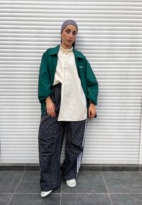 Casaco verde oversized, camisa creme e calças largas pretas com padrão de riscas brancas. Os acessórios incluem joias douradas e ténis de cor clara.