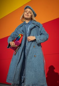 Τζιν καμπαρντίνα με μεγάλα πέτα, κούμπωμα με κουμπιά και ταιριαστό μπερέ. Κρατάει μια πολύχρωμη τσάντα ενάντια σε έντονο κόκκινο και πορτοκαλί φόντο.