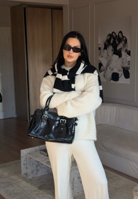 Oversized cream sweater with a textured surface, black and white striped scarf, black leather handbag, and white wide-leg trousers.
