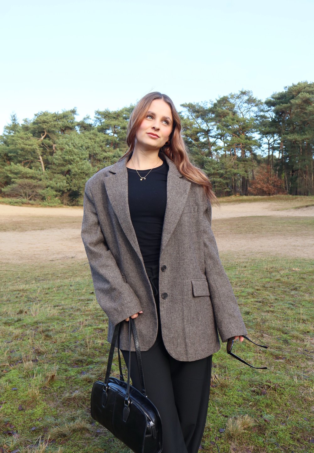 Blazer in lana grigia oversize con due bottoni, tasca frontale, abbinato a una maglietta nera e a una borsa nera, ambientato in un contesto naturale all'aperto.