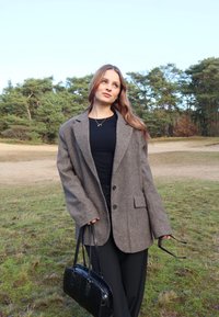 Blazer in lana grigia oversize con due bottoni, tasca frontale, abbinato a una maglietta nera e a una borsa nera, ambientato in un contesto naturale all'aperto.