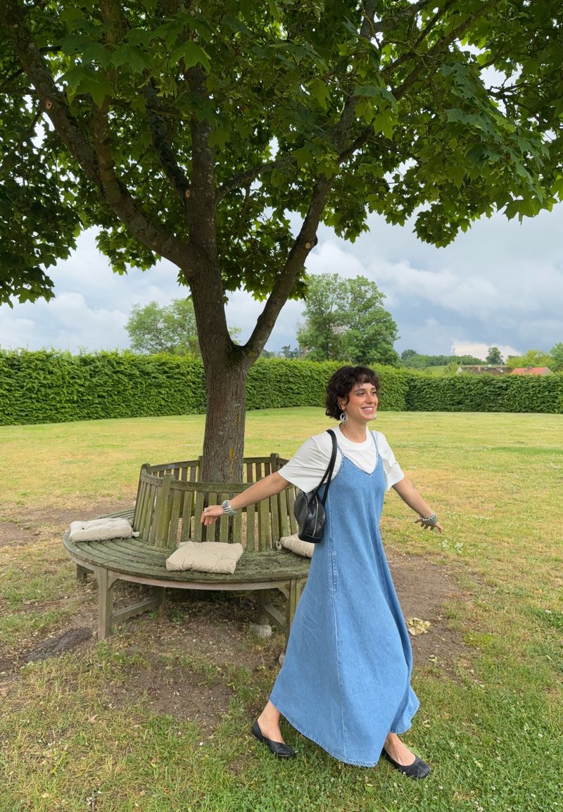 Femme portant une robe bleue et une chemise blanche tournant près d’un banc en bois circulaire autour d’un arbre dans un parc herbeux par une journée nuageuse.