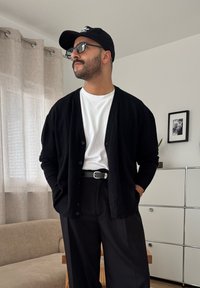 Férfi fekete sapkában, szemüveggel, fekete kardigán fehér ing felett, fekete nadrág övvel, modern nappaliban áll, kanapéval és szekrénnyel.