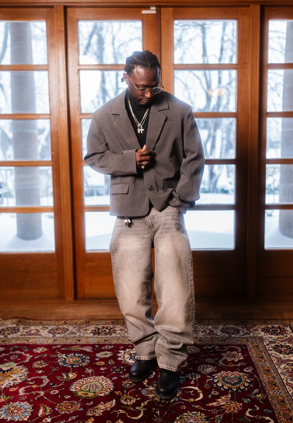 Vīrietis ar pītām matu lokām, valkājot brilles, slāņainas kaklarotas, pelēku žaketi, vaļīgas izbalējušas džinsas un melnus zābakus, stāv iekštelpās uz rakstaina paklāja.