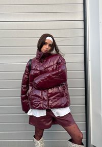 Femme portant une doudoune brillante bordeaux, un short en cuir assorti, des collants à pois, des bottes en peau de serpent et un bandeau blanc devant un mur gris.