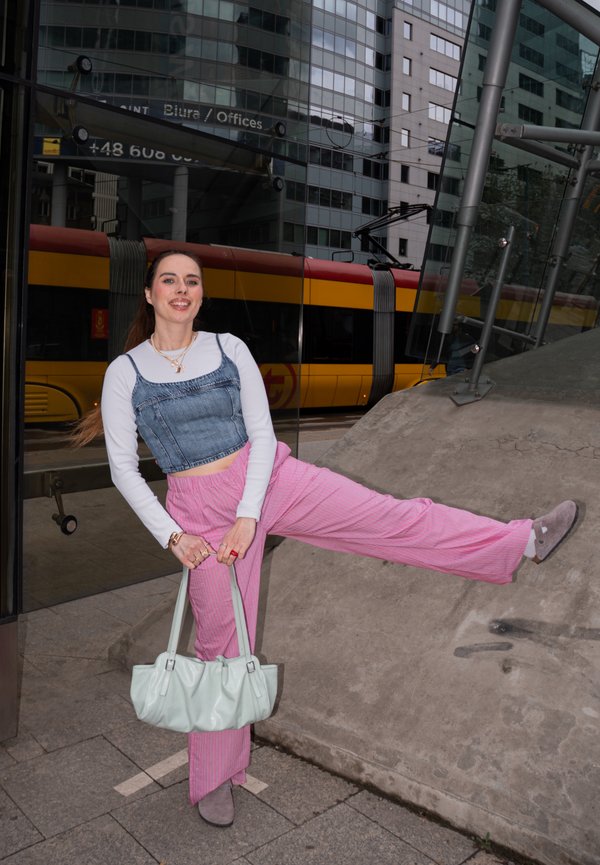Mujer con pantalones rosas y top vaquero posa en la acera de la ciudad, sosteniendo un bolso blanco, con un tranvía amarillo y edificios de vidrio al fondo.
