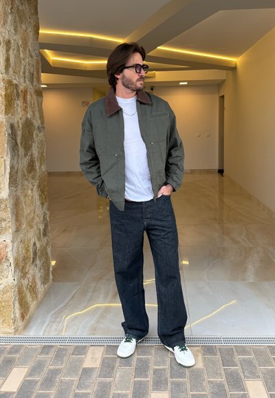 Chaqueta bomber verde oliva con cuello marrón, camiseta blanca, pantalones vaqueros anchos de mezclilla oscura y zapatillas blancas con cordones verdes.