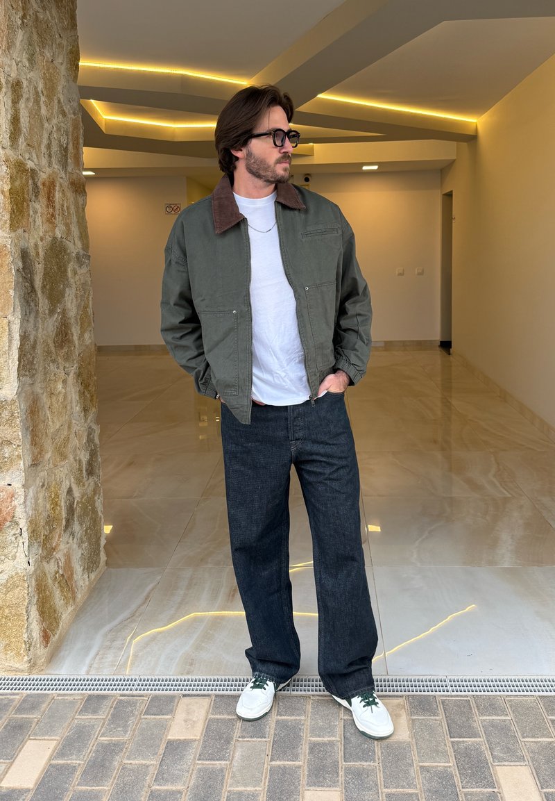 Olive green bomber jacket with brown collar, white t-shirt, dark denim wide-leg trousers, and white trainers with green laces.