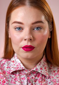 Close-up portret van een persoon met lang, kastanjebruin haar, die een bloempatroon shirt en opvallende magenta lipstick draagt tegen een roze achtergrond.