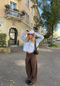 Młoda kobieta poprawiająca niebieski kaptur, ubrana w luźne brązowe spodnie i czarne buty, stojąca na miejskiej ulicy z drzewami i budynkami mieszkalnymi.