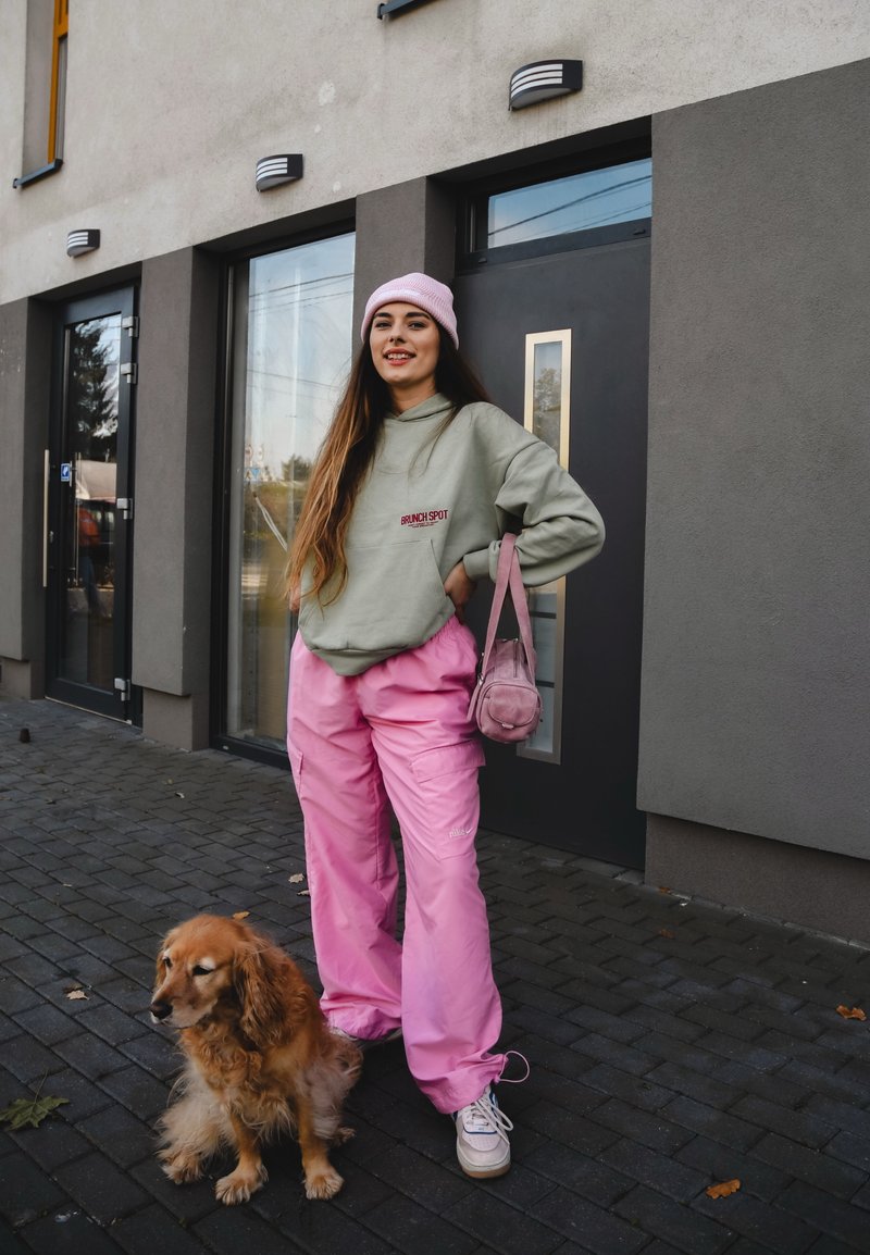 Μαρέν φούτερ με λογότυπο, ροζ cargo παντελόνι, λευκά αθλητικά παπούτσια, και μια μικρή ροζ τσάντα. Στεκόμενη δίπλα σε έναν χρυσό retriever σε κυβόλιθους.