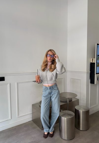 Joven mujer con ropa informal sostiene una bebida con hielo y posa ajustándose las gafas en una cafetería moderna con taburetes de metal y paredes blancas con paneles.