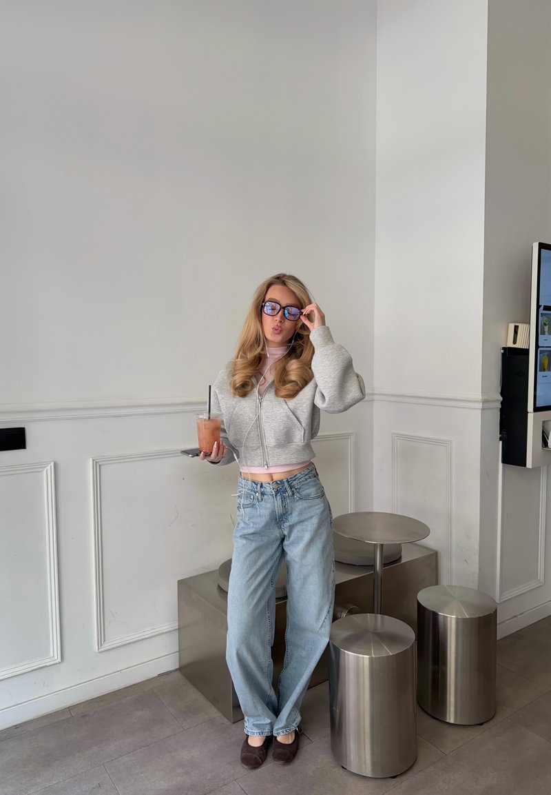 Νεαρή γυναίκα με casual ρούχα κρατά παγωμένο ποτό και ποζάρει διορθώνοντας τα γυαλιά της σε μοντέρνο καφέ με μεταλλικά σκαμπό και λευκούς τοίχους με πάνελ.