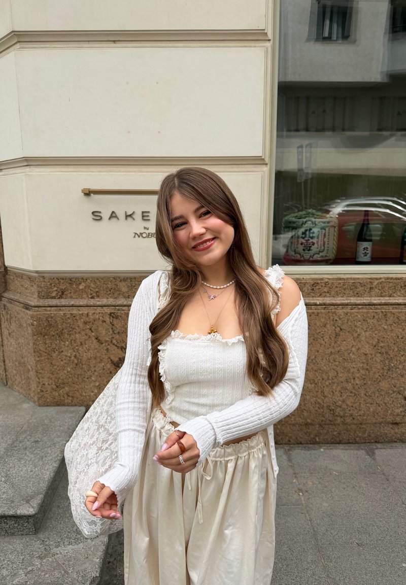 Jeune femme aux longs cheveux bruns portant un haut blanc et un pantalon fluide crème, sourit devant un bâtiment avec un panneau « SAKE ».