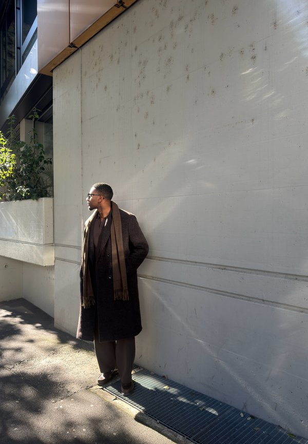 Hombre con gafas, abrigo largo oscuro y bufanda marrón, de pie en la acera junto a una pared de concreto gris claro, mirando hacia la izquierda.