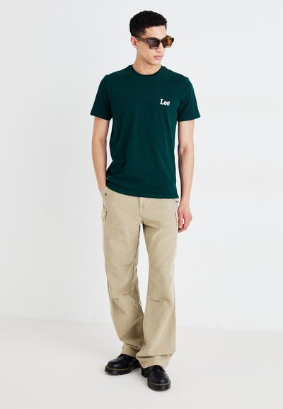 Camiseta de algodón verde con el logo blanco de "Lee", combinada con pantalones cargo beige con bolsillos y zapatos negros gruesos. Modelo con gafas de sol de gran tamaño.