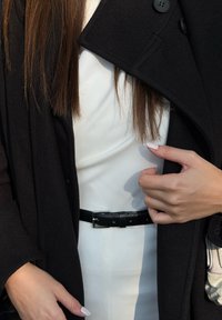 Cojoc negru din lână cu reveruri mari, închis cu un nasture; rochie albă dedesubt, strânsă în talie cu o curea neagră din piele.