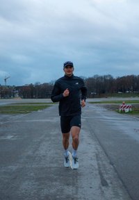 Sportists skrien pa mitrai asfaltam, ģērbies melnā jakā un šortos, ar baltām kedām un zilu cepuri, zem mākoņainas debess.
