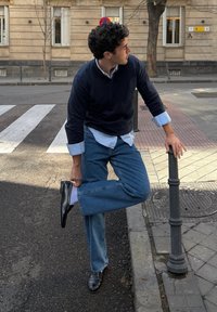 Άνδρας με μπλε τζιν και μαύρα παπούτσια στέκεται στο δρόμο, κρατώντας το πόδι πίσω ενώ στηρίζεται σε μαύρο πασσάλό κοντά σε διάβαση πεζών.