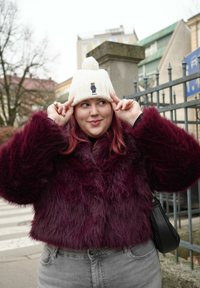 Γυναίκα που προσαρμόζει ένα λευκό πλεκτό καπέλο με σχέδιο αρκούδας, φορώντας μπορντό παλτό από συνθετική γούνα και γκρι τζιν, στέκεται έξω κοντά σε μεταλλικό φράχτη.