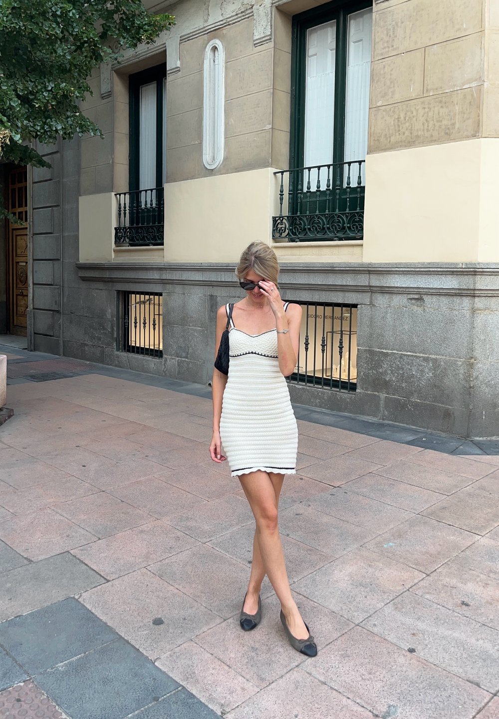 Λευκό πλεκτό φόρεμα με μαύρες λεπτομέρειες, με λεπτές τιράντες και τελείωμα με κυματιστό σχέδιο. Φορείται με μαύρες φλατ παντόφλες, σε φόντο από πεζοδρόμιο και κτίριο από πέτρα.