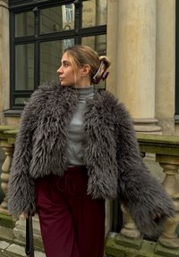 Φουντωτό γκρι ψεύτικο γούνινο μπουφάν με κροπαρισμένο σχέδιο, φορεμένο πάνω από ένα ανοιχτό γκρι φουλάρι και σε συνδυασμό με μπορντό παντελόνι.