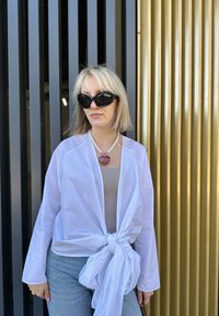 Un wrap blanc léger avec un nœud à l'avant, porté sur un haut beige et associé à un jean. Comprend un collier en forme de cœur et des lunettes de soleil noires.