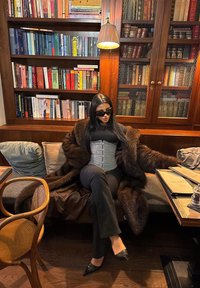 Γυναίκα με γυαλιά ηλίου, φορώντας γούνινο παλτό και κορσέ, καθισμένη σε μαξιλαροκαθισμένο πάγκο σε βιβλιοθήκη με ξύλινη επένδυση, με ράφια γεμάτα βιβλία και φωτιστικό από πάνω.