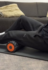 Pantaloni sportivi neri con texture e logo Puma, posizionati sopra un rullo di schiuma grigio con un accento arancione, su un tappetino da esercizio con texture.
