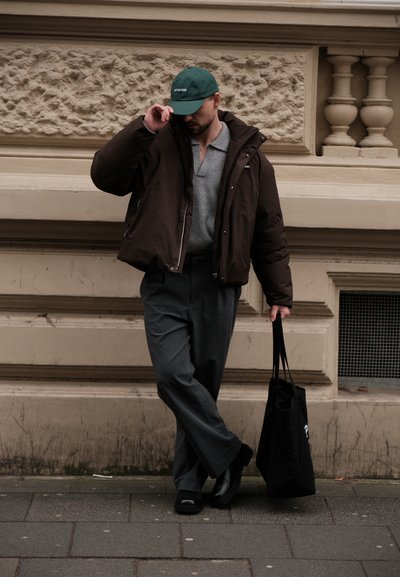 Chaqueta acolchada marrón, camiseta de punto gris, pantalones de pierna ancha grises, zapatos negros, gorra verde con texto, bolsa negra, de pie en una calle de adoquines.