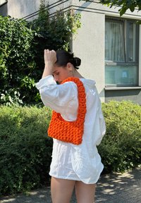 Πορτοκαλί πλεκτή τσάντα με χοντρή υφή και φαρδύ λουρί, συνδυασμένη με μια χαλαρή λευκή μπλούζα σε εξωτερικό χώρο με πράσινα φυτά.