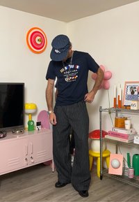Tricou bleumarin cu design grafic, pantaloni largi cu dungi, șapcă gri și pantofi negri. Accesoriile includ coliere straturi și o brățară.
