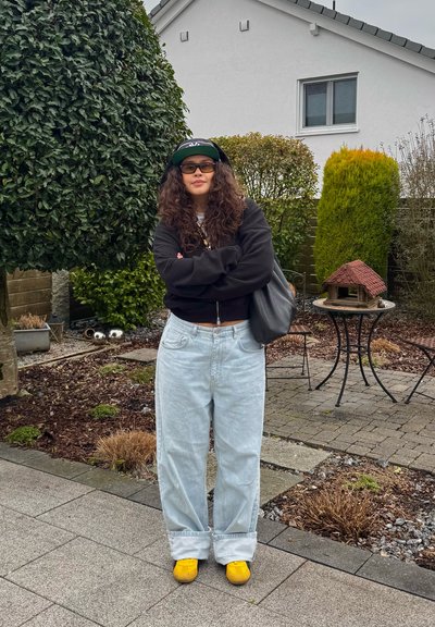 Persona joven con cabello largo y rizado, vestida con sudadera negra con capucha, gorra, gafas, jeans anchos claros y zapatillas amarillas, parada en un jardín con arbustos y una mesa pequeña.